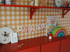 Ockergelbe Gingham-Tapete von Atelier Chien mit rotem Wandregal und Schrank, in Nahaufnahme.